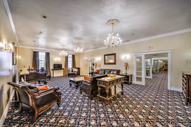 A spacious, elegantly furnished living room with patterned carpet, multiple chandeliers, leather armchairs and sofas, wooden tables, lamps, and a television near large windows with curtains. The room has light-colored walls and ceiling with decorative molding, and a doorway leading to a hallway.
