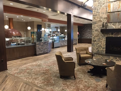 Interior view of a senior living facility dining area with a serving counter and food display on the left side. There are three upholstered armchairs arranged around a round black coffee table on a patterned area rug. A stone fireplace with decorative items is visible on the right side.