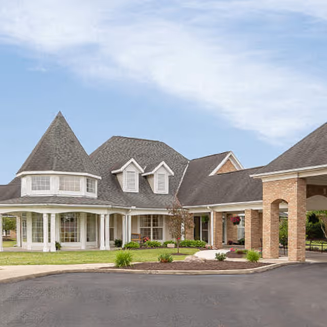 Exterior view of a senior living facility with a large, multi-gabled roof, a turret, and a covered entrance supported by brick columns. The building is surrounded by a landscaped area with grass, shrubs, and a paved driveway under a partly cloudy sky.