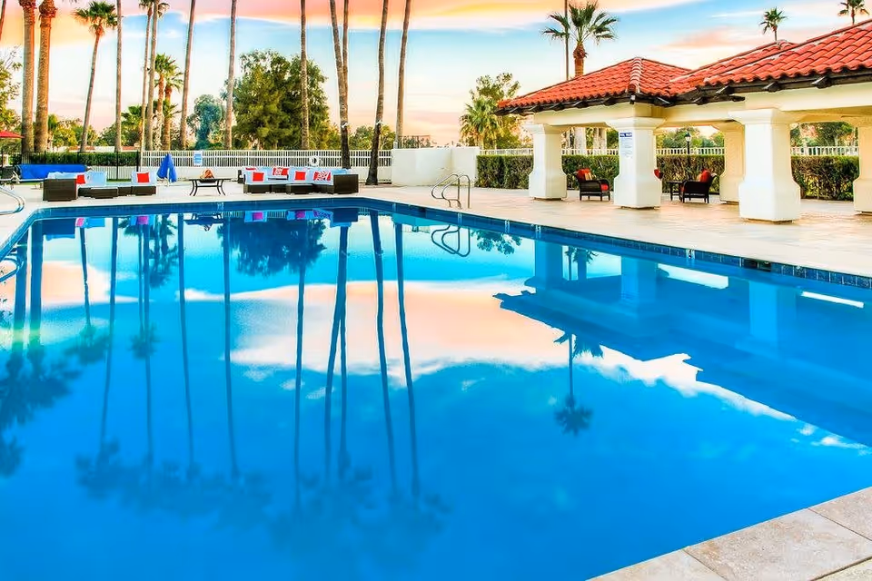 Outdoor swimming pool at a senior living facility with palm trees reflected in the water, lounge chairs with red and white cushions, and a covered seating area with a red-tiled roof under a colorful sunset sky.