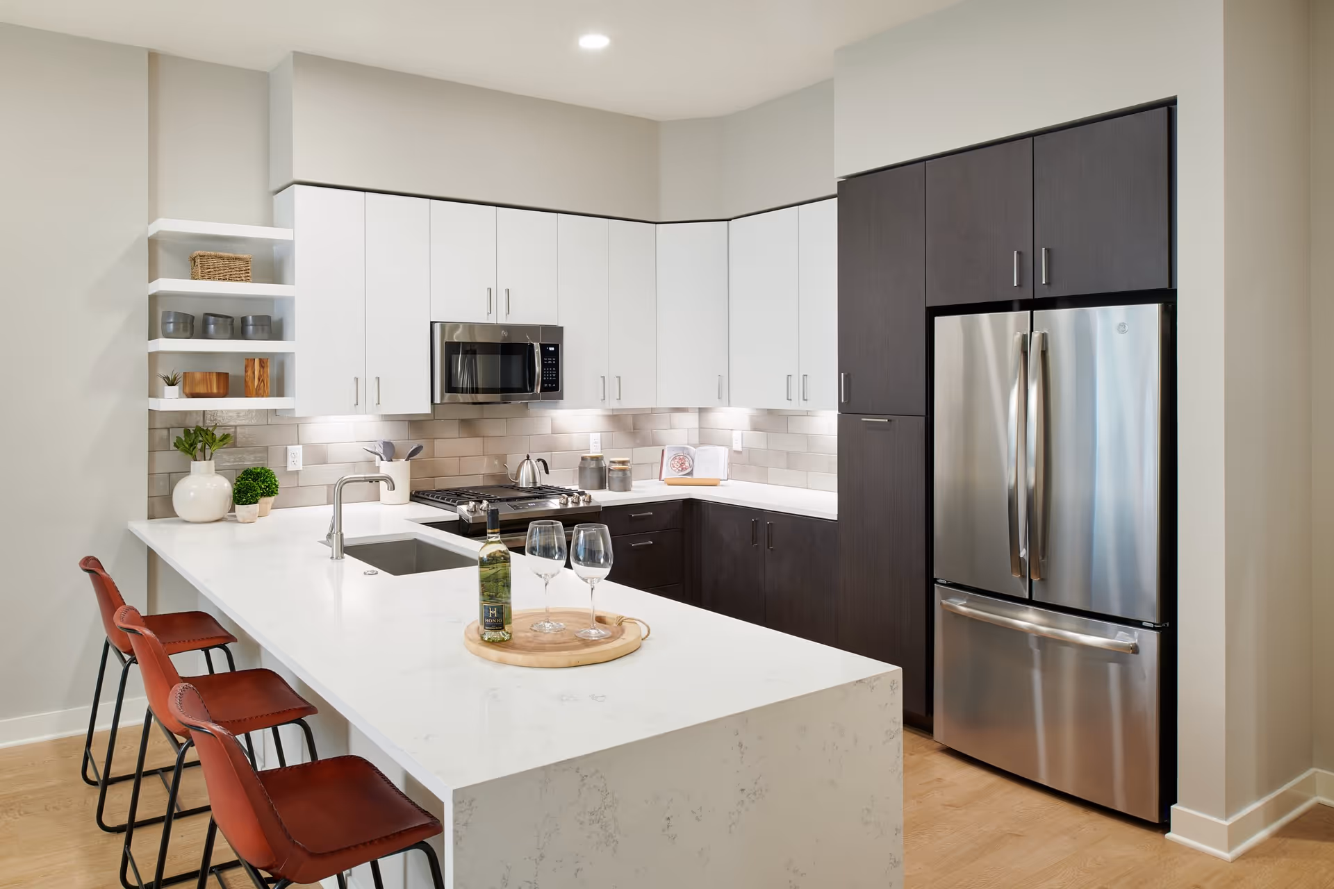 Modern open kitchen with a white island and sink, three red bar stools, stainless steel refrigerator and microwave, and white and dark cabinetry.