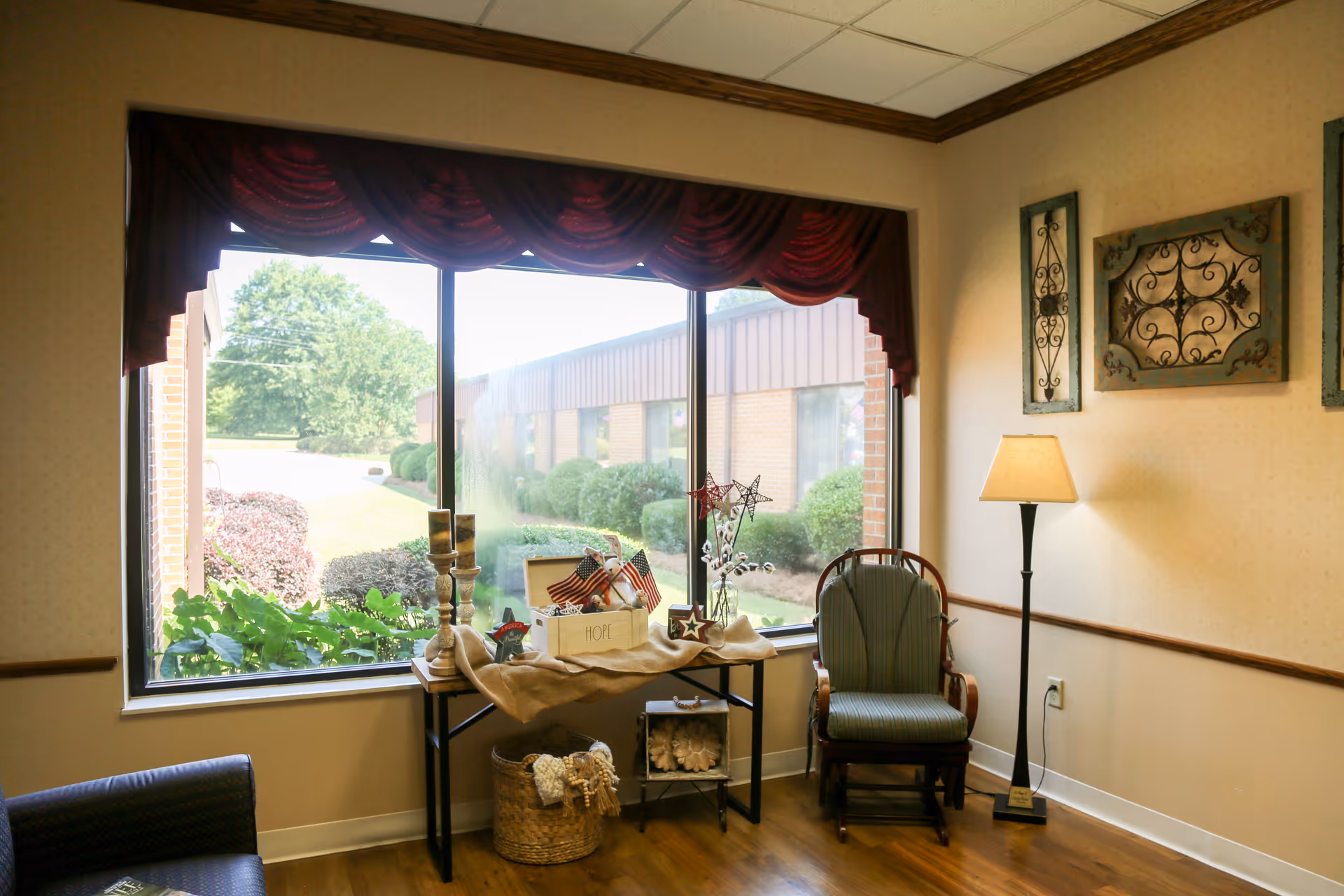 A cozy sitting area in a senior living facility with a large window showing outdoor greenery. The room features a cushioned wooden chair, a floor lamp, a small table decorated with patriotic items and candles, and wall art with wrought iron designs.