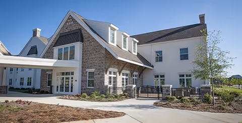 Exterior view of a large, modern building with a combination of stone and white siding. The building has multiple windows, a covered entrance, and a paved driveway with landscaped areas including small plants and trees under a clear blue sky.