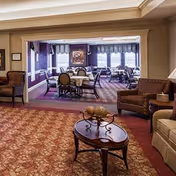 A cozy interior common area with patterned carpet, a wooden coffee table with decorative items, armchairs, and a view into a dining room with multiple tables and chairs near windows with curtains.