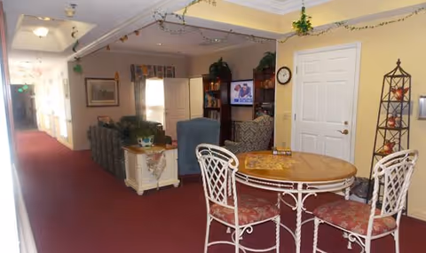 Interior view of a senior living facility common area with a round table and four chairs in the foreground, a seating area with armchairs and a couch, a television mounted on the wall, and decorative plants and artwork. The space has carpeted floors and light-colored walls with ceiling lights.
