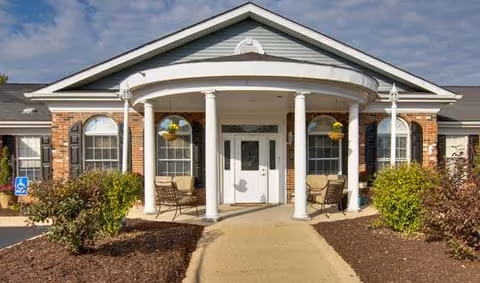 Front exterior view of a single-story brick building with a covered entrance supported by four white columns. There are two chairs and hanging flower baskets on the porch. The sky is partly cloudy and there are bushes and a paved walkway leading to the entrance.