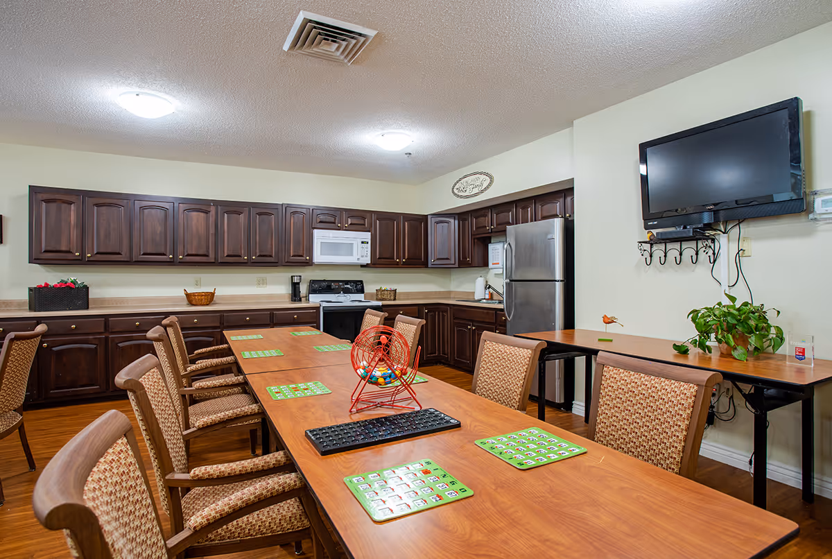 A communal room with a long wooden table surrounded by cushioned chairs. On the table are bingo cards and a red bingo cage with colorful balls. The room has a kitchen area with dark wooden cabinets, a white microwave, a stove, and a stainless steel refrigerator. A flat-screen TV is mounted on the wall above a small table with a green plant.