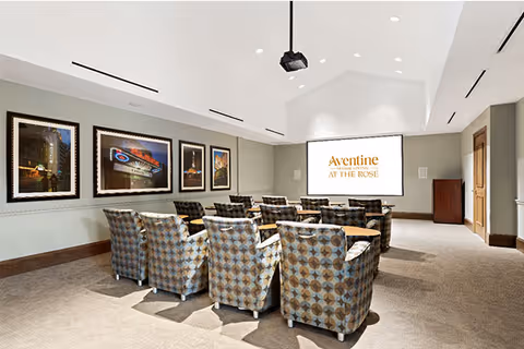 Small theater-style interior with patterned armchairs arranged facing a ceiling-mounted projector and a screen displaying 'Aventine at the Rose', with framed artwork on the side wall.