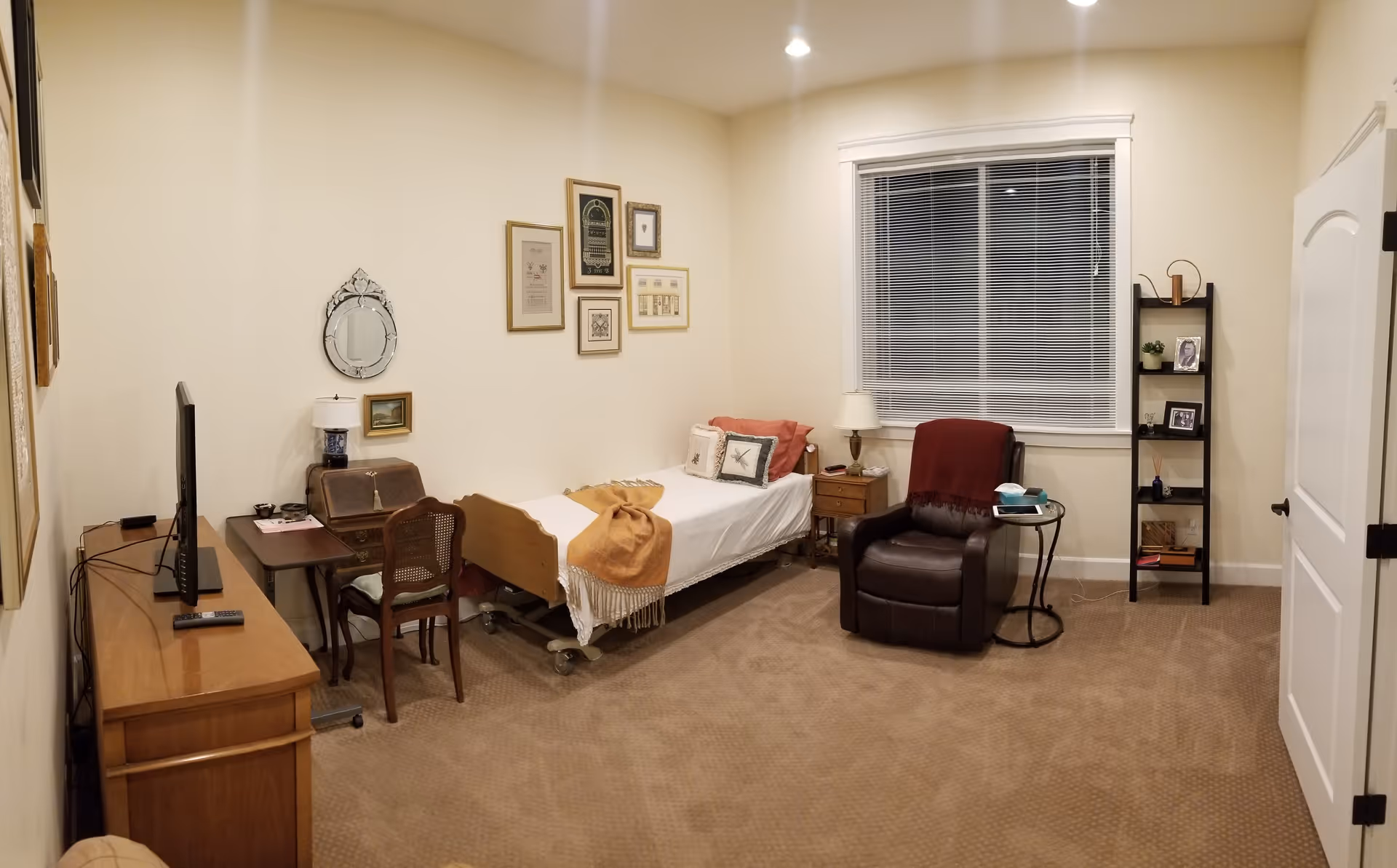 A cozy bedroom in an elderly care facility featuring a single bed with white bedding and decorative pillows, a brown recliner chair with a red throw blanket, a small wooden nightstand with a lamp, a wooden desk with a chair and a TV on a dresser, framed pictures and a mirror on the wall, a window with blinds, and a black shelving unit with personal items.
