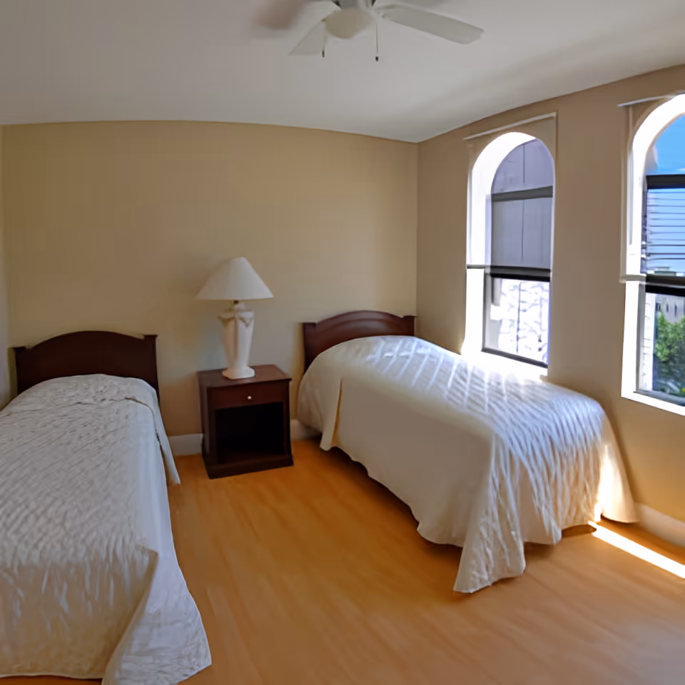 A bedroom with two single beds covered in white bedspreads, a wooden nightstand with a white lamp between the beds, light wood flooring, beige walls, and two arched windows letting in natural light.
