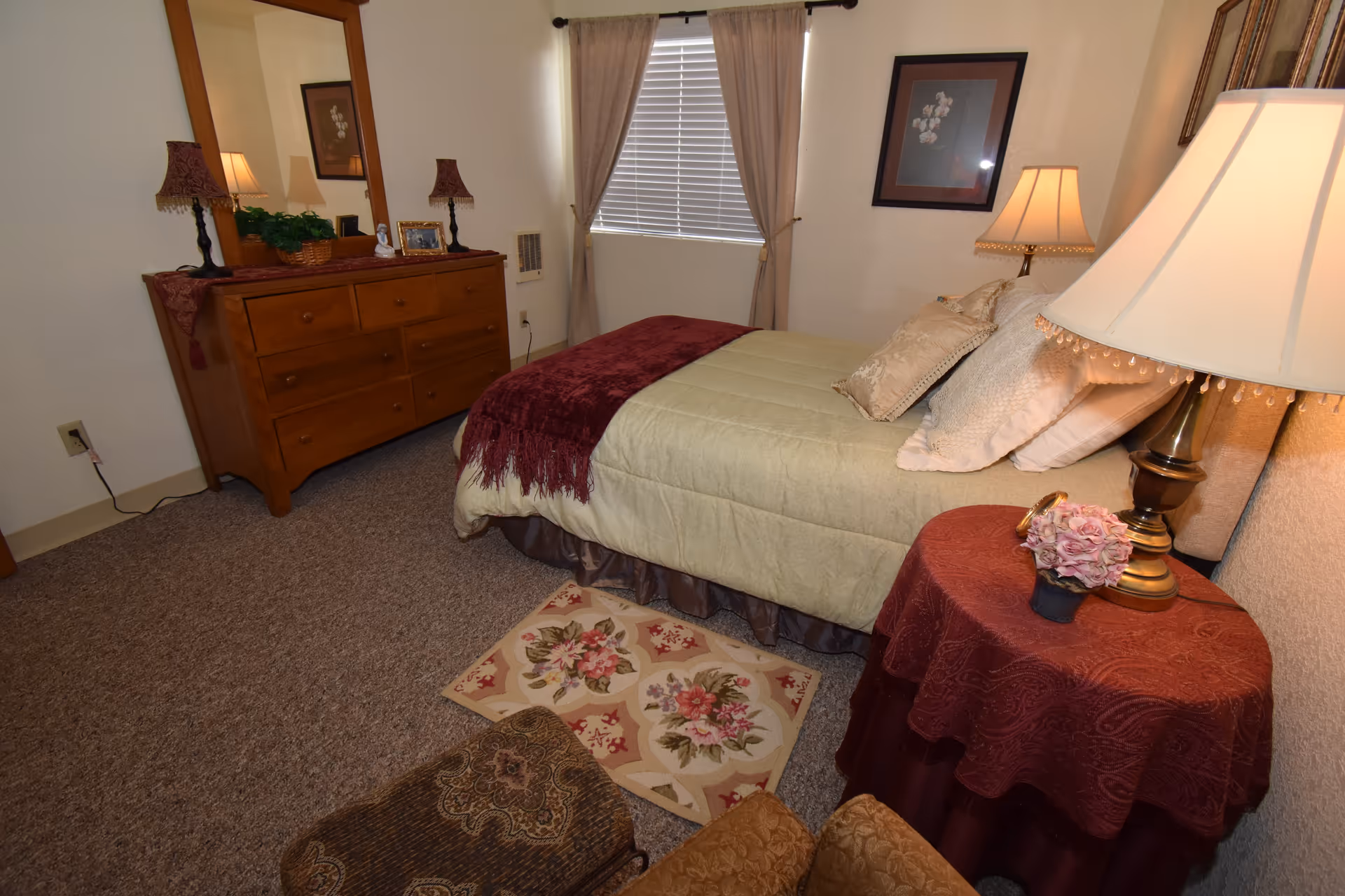 A cozy bedroom with a bed covered in beige and burgundy bedding, a wooden dresser with a large mirror, two table lamps, a small round table with a burgundy tablecloth, and a floral rug on the carpeted floor. There is a window with beige curtains and a framed picture on the wall.