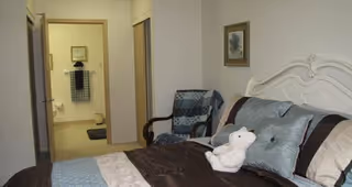 A bedroom with a neatly made bed featuring decorative pillows and a white teddy bear. There is a wooden chair with a blue and gray quilt draped over it. The room has light-colored walls and an open doorway leading to a bathroom with towels hanging on the wall.
