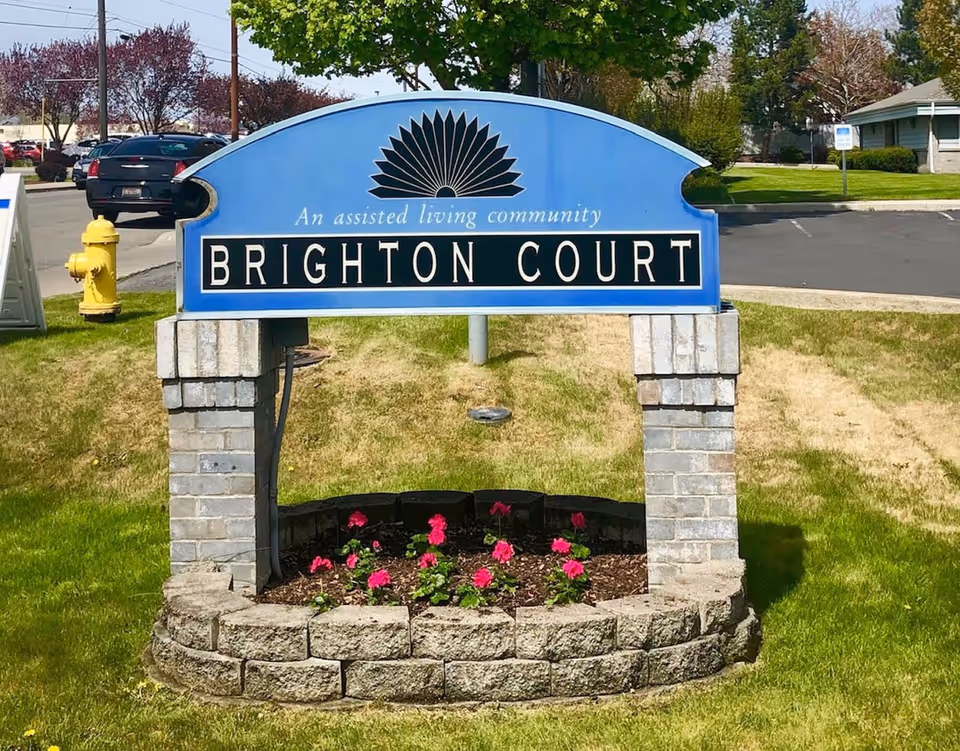 A blue sign for Brighton Court, an assisted living community, mounted on two brick pillars with a small flower bed of pink flowers at the base. The sign is located on a grassy area near a parking lot with cars and trees in the background.