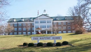 Front exterior view of a large three-story brick building with a green roof and a central cupola, set on a grassy hill with a sign in front that reads 'THE BAPTIST HOME'. Leafless trees are visible on either side of the building under a clear sky.