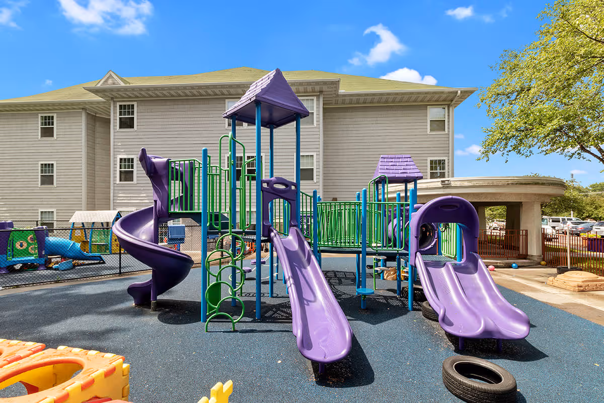 Outdoor playground area with purple slides, climbing structures, and green railings in front of a multi-story beige building under a blue sky with some clouds.