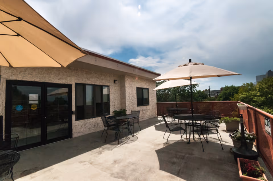 Outdoor patio area with several round metal tables and chairs, each shaded by large beige umbrellas. The patio is adjacent to a building with a textured exterior wall and several windows. There are potted plants along the railing and a partly cloudy sky overhead.