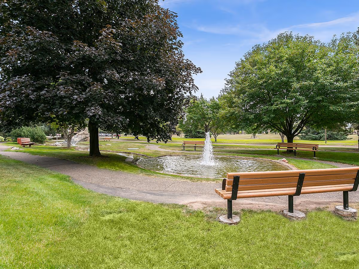 A peaceful outdoor park area with green grass, several large trees, a small pond with a water fountain in the center, and multiple wooden benches along a paved walking path.