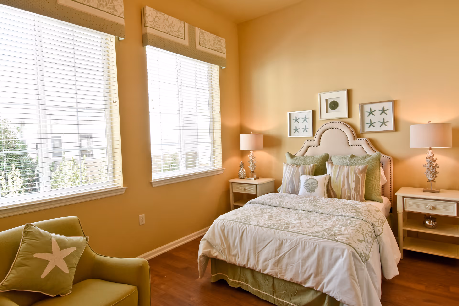 A cozy bedroom with a neatly made bed featuring multiple pillows and a patterned bedspread. The room has warm beige walls, two large windows with white blinds, two matching bedside tables with lamps, and framed artwork above the bed. There is also a green armchair with a starfish pillow near the windows.