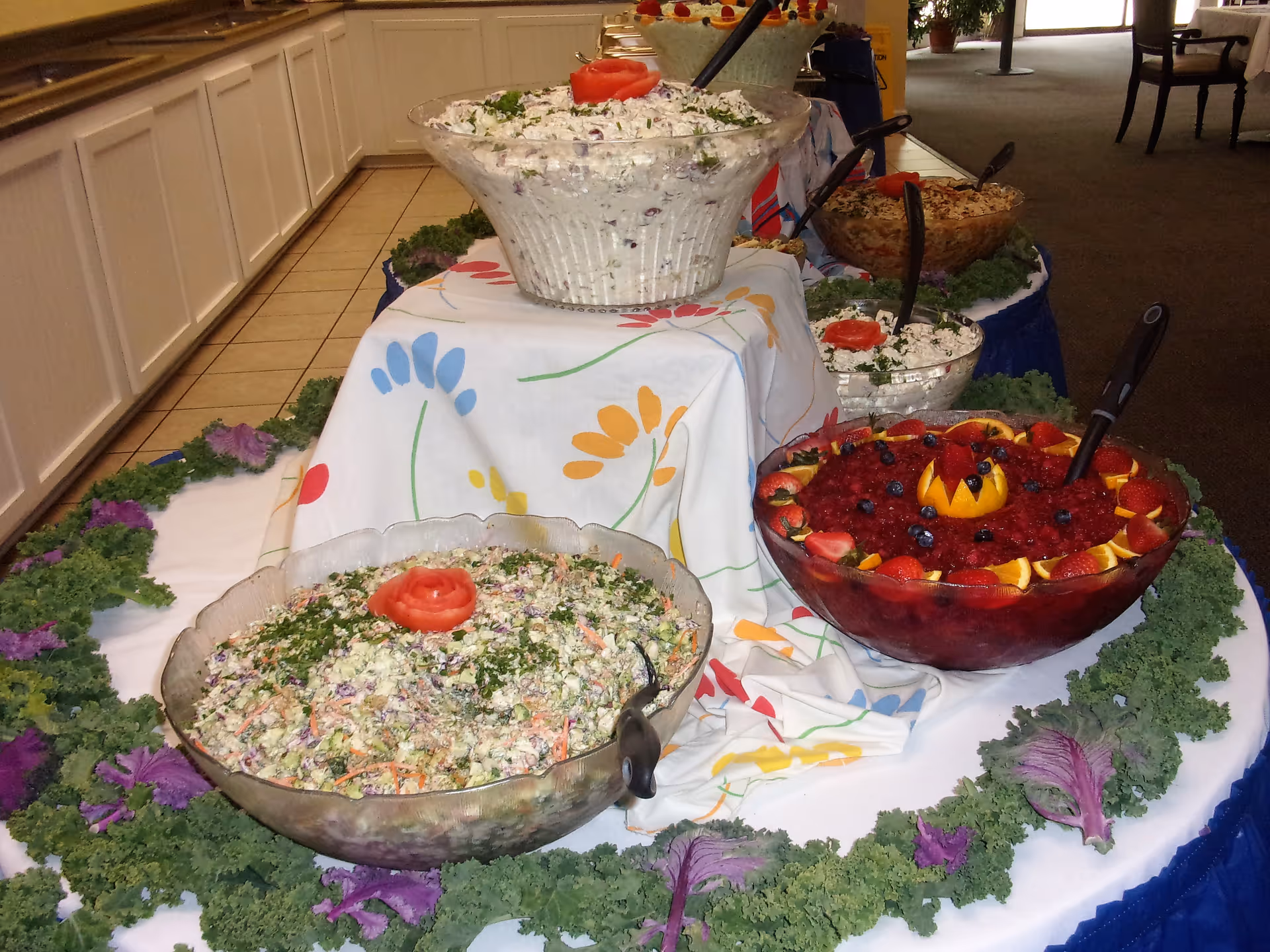 A buffet table with several large bowls of various salads and fruit punch, decorated with leafy greens and a colorful floral tablecloth, set in an indoor dining area.