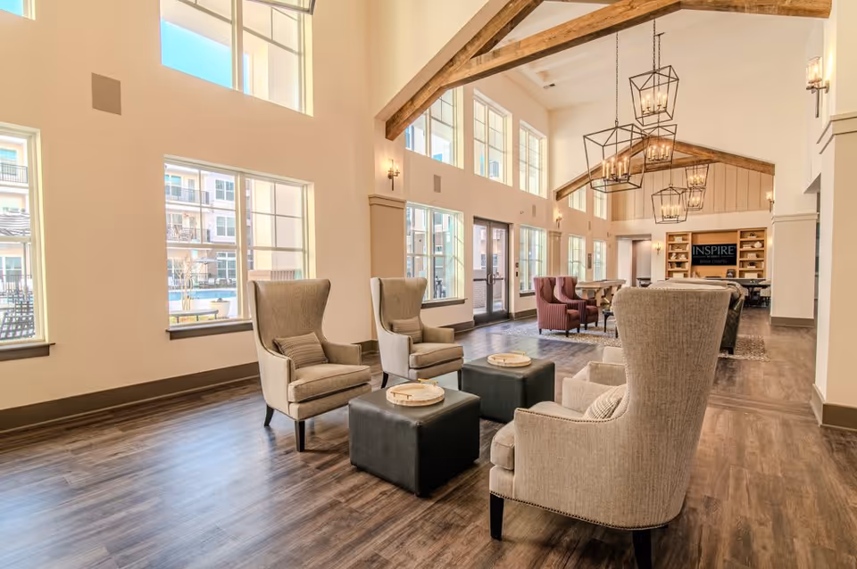 A spacious and well-lit living room area in Inspire Briar Chapel featuring high ceilings with exposed wooden beams and large windows allowing natural light to fill the space. The room is furnished with beige wingback chairs arranged around black ottomans, and additional seating including red armchairs and a dining table in the background. Modern pendant lights hang from the ceiling, and a built-in shelving unit with a TV displaying the Inspire Briar Chapel logo is visible at the far end.