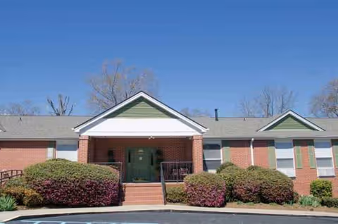 Brick senior living facility entrance with green doors, steps, shrubs, and a clear blue sky.