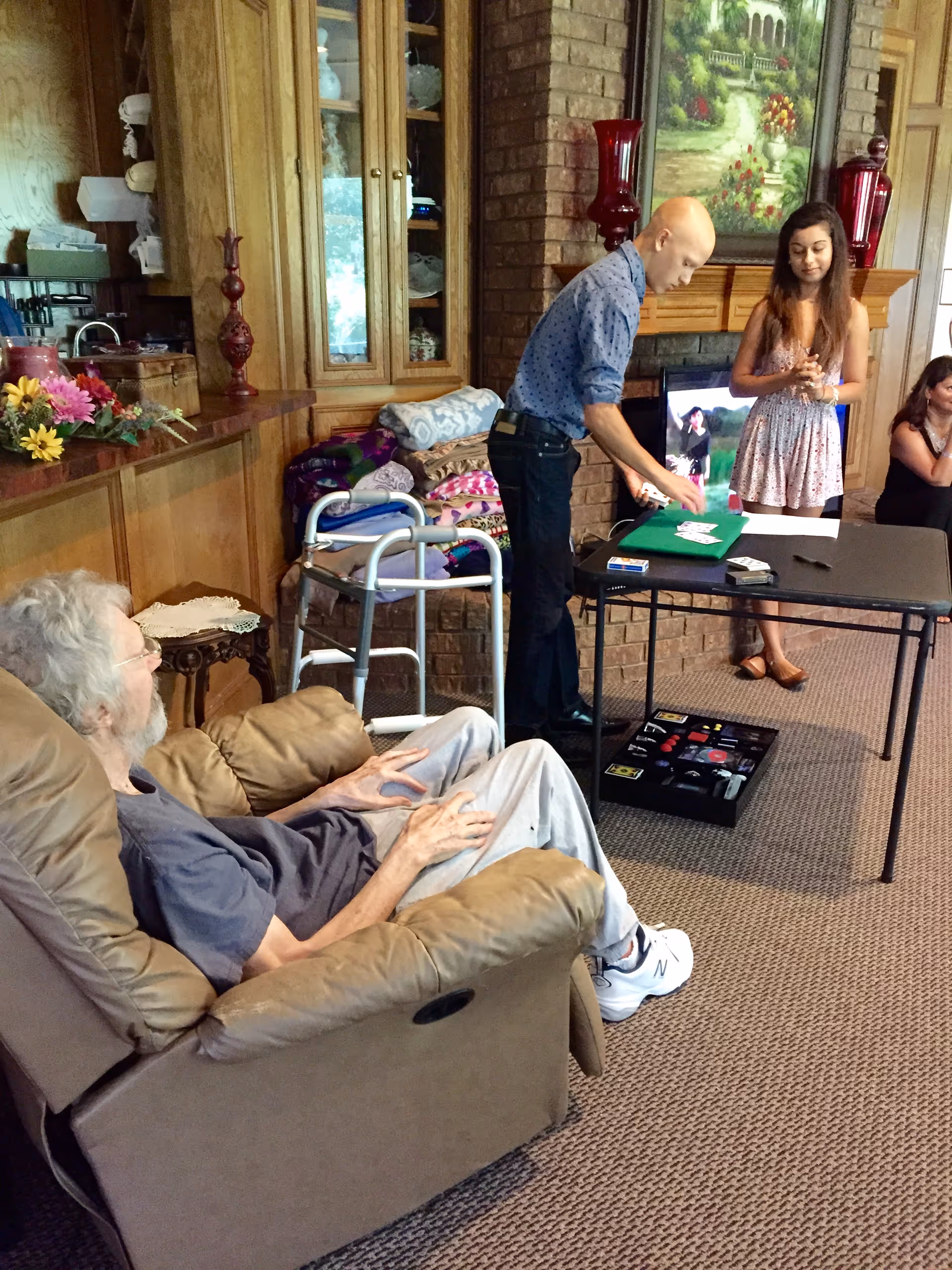 An elderly man sits in a beige recliner chair in a cozy living room with wooden cabinets and a brick fireplace. A young man is performing a card trick on a folding table while two women watch. The room has a carpeted floor and various decorative items including flowers and a painting above the fireplace.