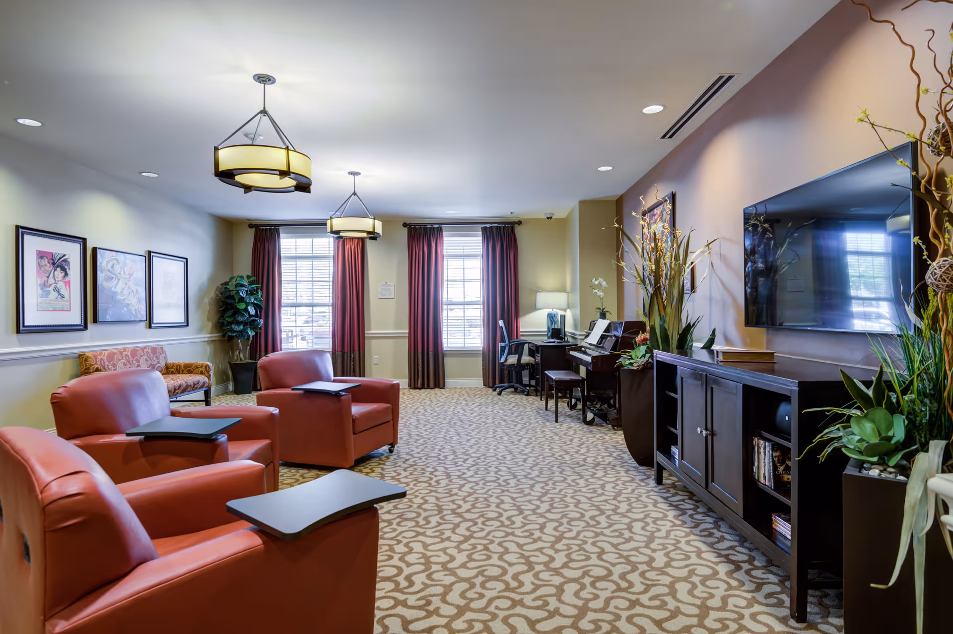 A cozy living room area with three red armchairs each equipped with a small black tray table. The room has patterned carpet flooring, two large windows with red curtains, and a piano with a chair near the windows. On the right side, there is a large flat-screen TV mounted on the wall above a dark wooden cabinet with plants and decorative items around it. The walls are adorned with framed artwork and the ceiling has two modern hanging light fixtures.