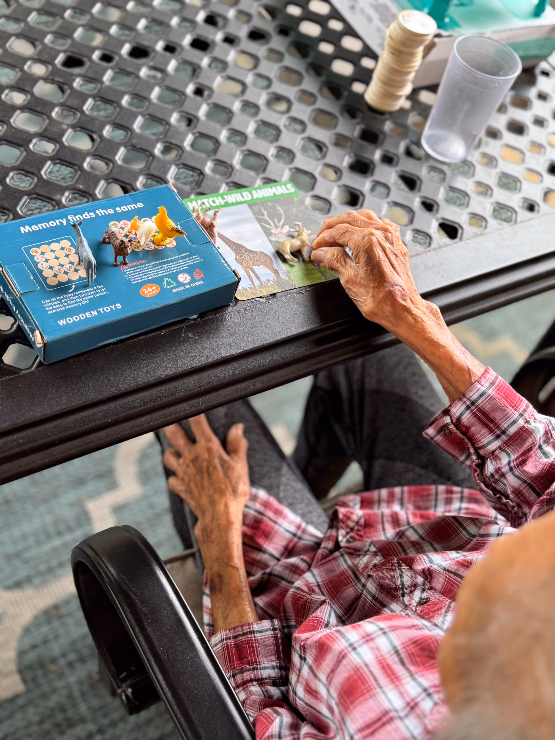 An elderly person wearing a red plaid shirt sits at a metal table playing a memory matching game with small animal figurines and cards labeled 'MATCH WILD ANIMALS'. The person's hands are visible as they interact with the game pieces on the table.