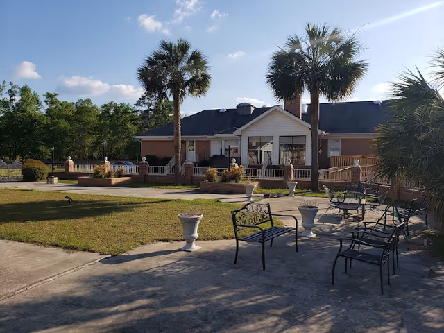 Outdoor patio area with several black metal benches and white decorative planters on a concrete surface. In the background, there is a single-story brick building with a white porch and railing, flanked by tall palm trees. The sky is clear with a few clouds, and there is greenery surrounding the area.
