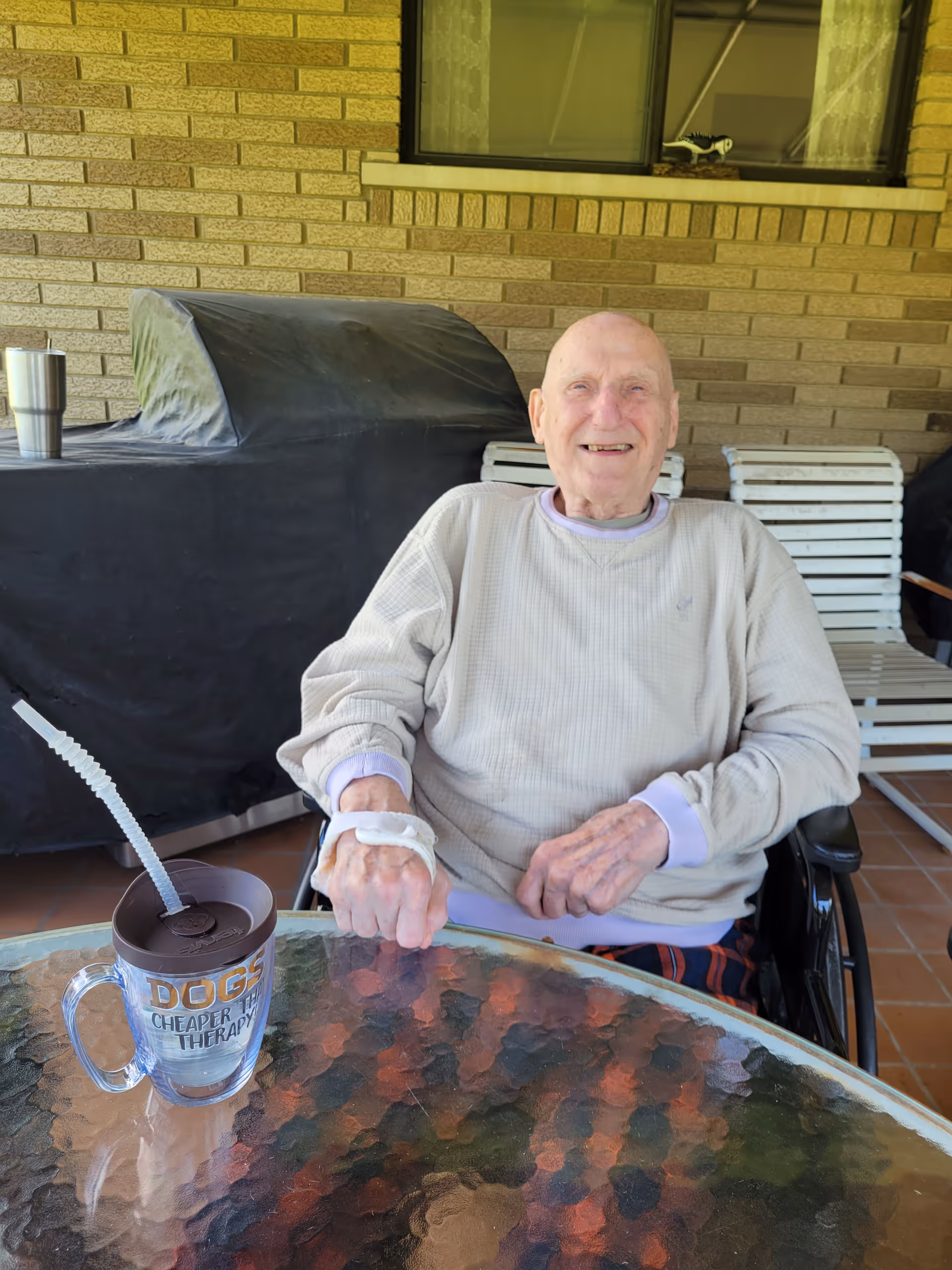 An elderly man sitting in a wheelchair at a glass table outdoors, smiling. He is wearing a light-colored sweater and has a medical bandage on his hand. On the table is a clear mug with a brown lid and straw that reads 'DOGS CHEAPER THAN THERAPY'. Behind him is a brick wall with a window and some white patio chairs.