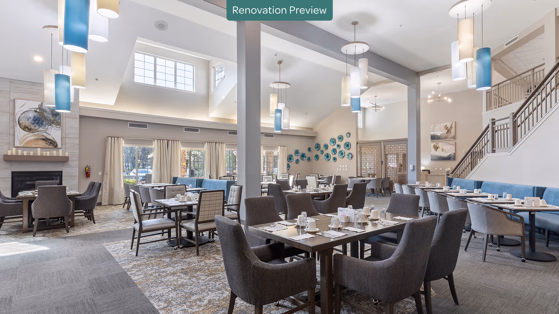 Spacious and well-lit dining room with multiple tables and chairs arranged neatly. The room features high ceilings with modern pendant lights, large windows with curtains allowing natural light, a fireplace with artwork above it, and decorative wall art. There is a staircase visible on the right side and a sign at the top center that reads 'Renovation Preview'.