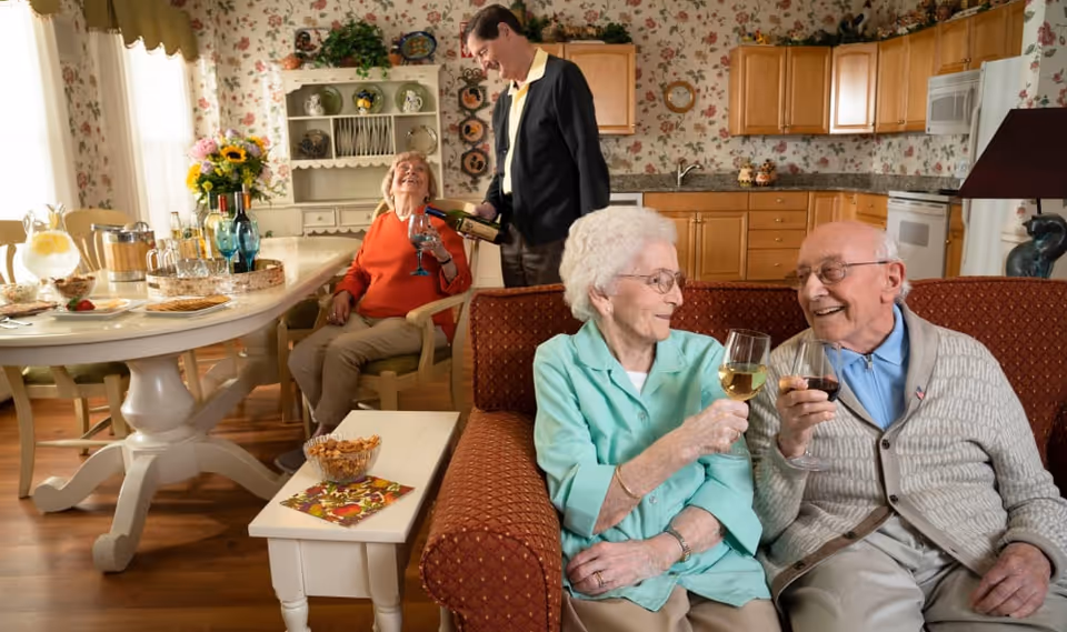 Two elderly people sit on a couch clinking wine glasses while another seated woman and a man pouring drinks are in the adjoining kitchen-dining area.