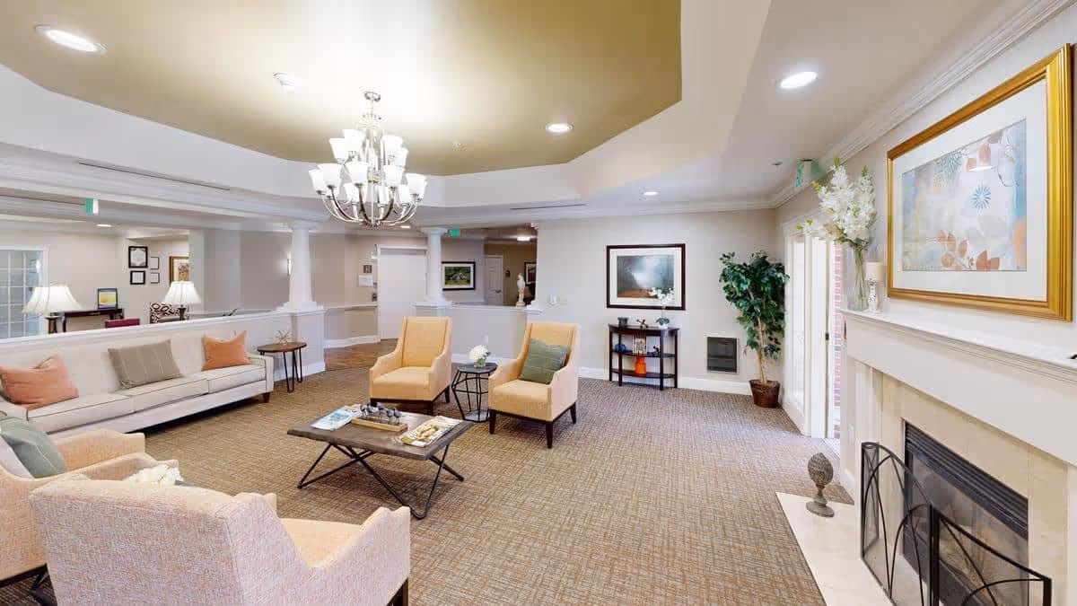 A spacious and well-lit living room area with beige carpeting, a white sofa with pink and beige cushions, two beige armchairs, a wooden coffee table with magazines, a chandelier hanging from the ceiling, a fireplace with a framed artwork above it, a potted plant, and a small table with decorative items.