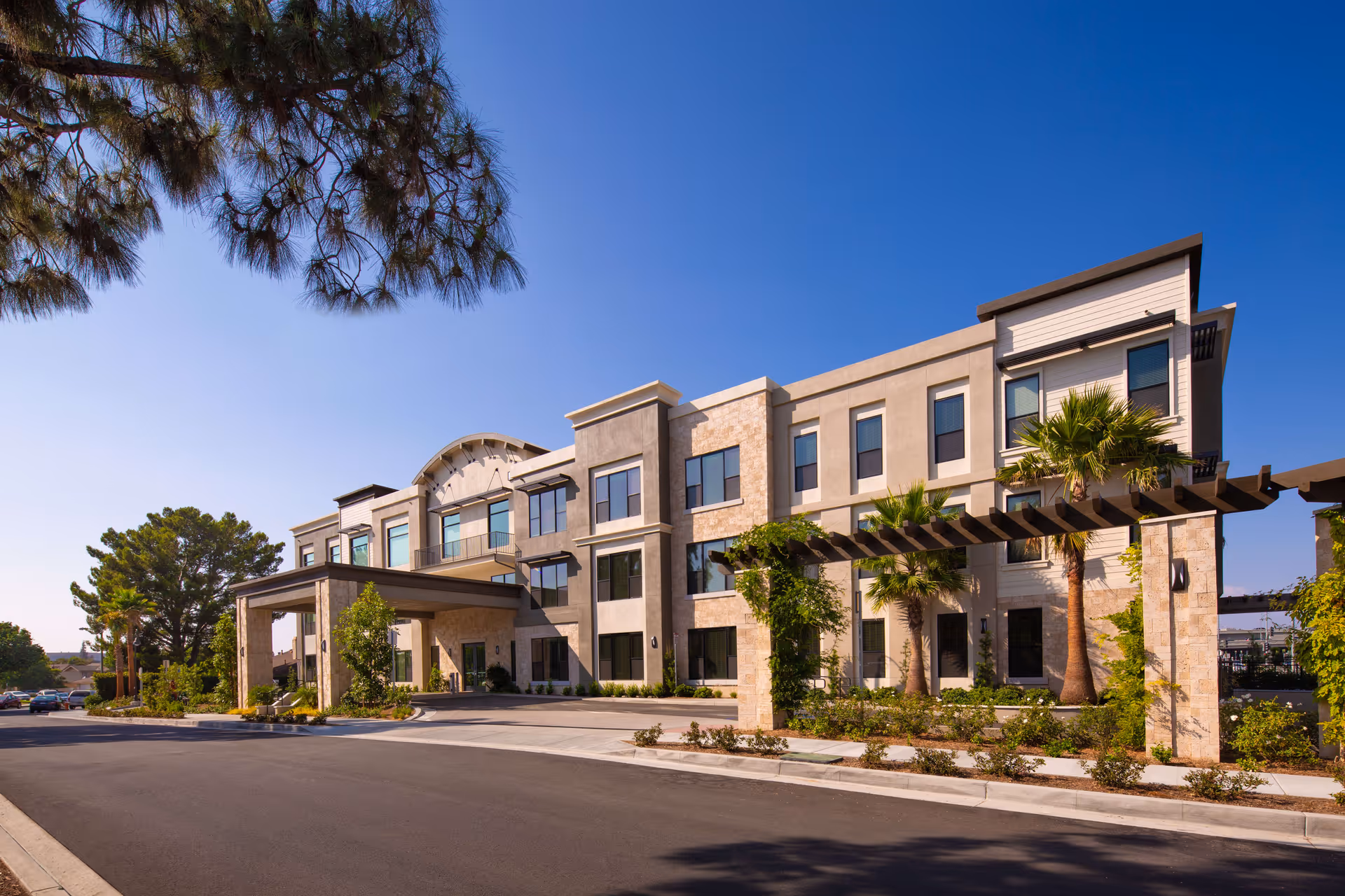 Exterior view of a modern three-story senior living facility building with large windows, a covered entrance, palm trees, and landscaped greenery under a clear blue sky.