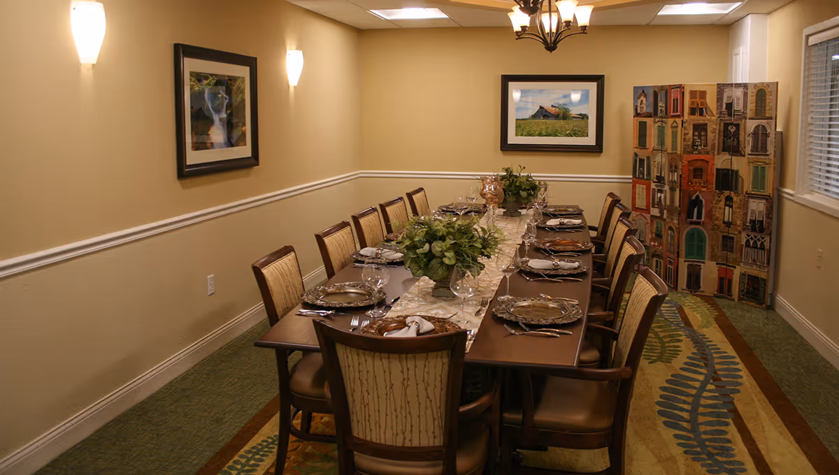 A long dining table set for a meal with plates, glasses, and silverware in a warmly lit room. The table has a decorative runner and green floral centerpieces. There are twelve upholstered chairs around the table. The walls are beige with white trim, decorated with framed pictures and a colorful folding screen near a window with blinds.