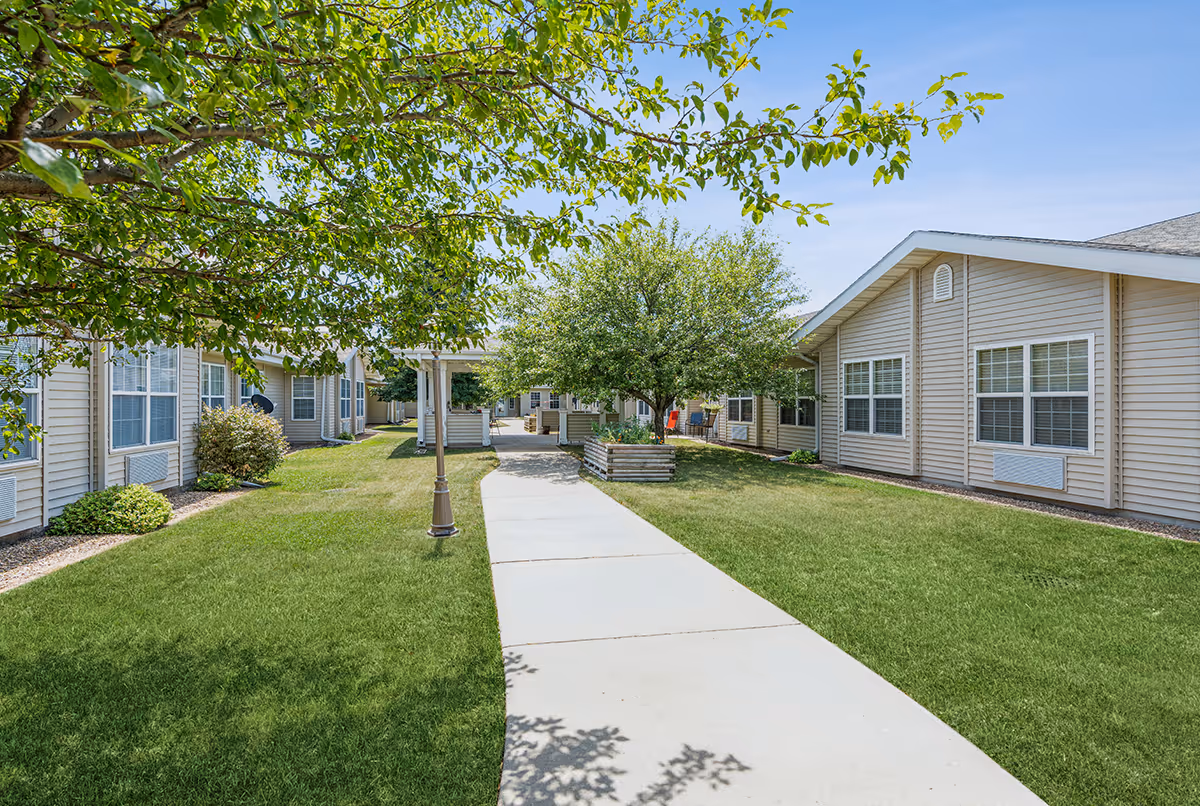 A concrete walkway through a grassy courtyard between single-story beige senior living buildings lined with trees and a lamppost.