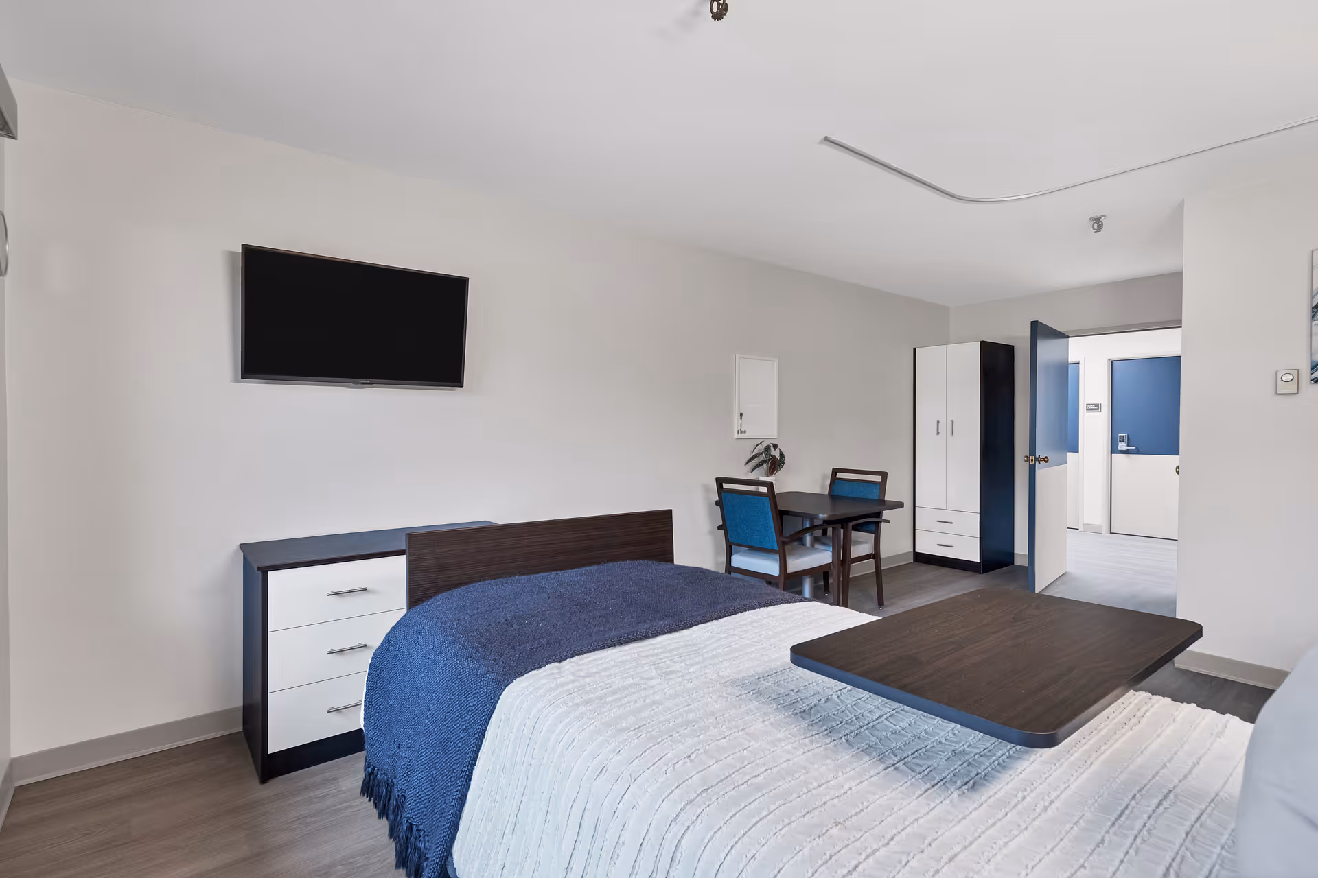 A clean and modern bedroom in a senior living facility with a bed covered in white and blue blankets, a mounted flat-screen TV on the wall, a small dresser, a table with two chairs, and a wardrobe. The room has light-colored walls and flooring, with an open door leading to a hallway.