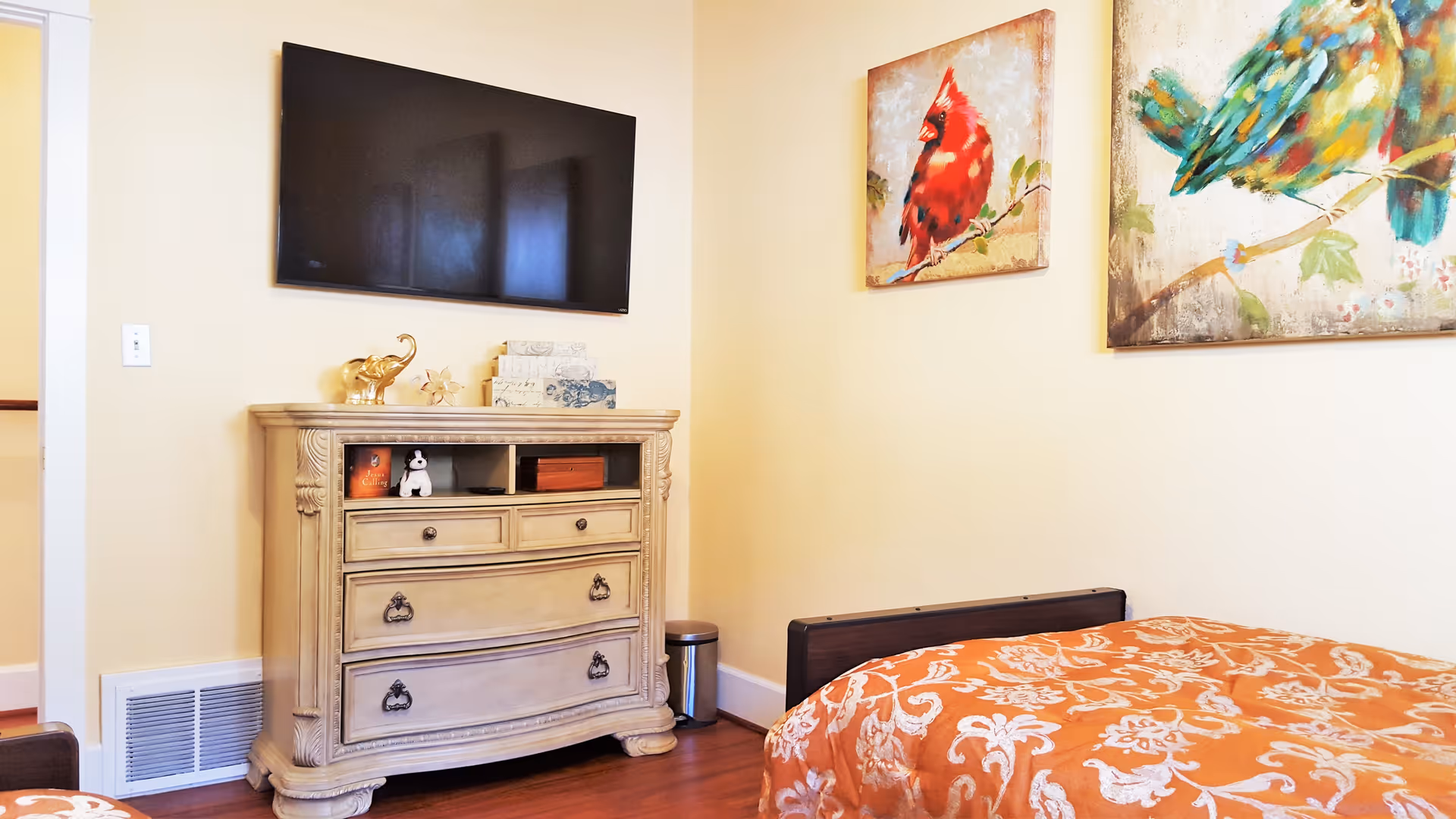 Bright bedroom with an ornate dresser under a wall-mounted TV, patterned orange bedspread, and bird artwork on the wall.