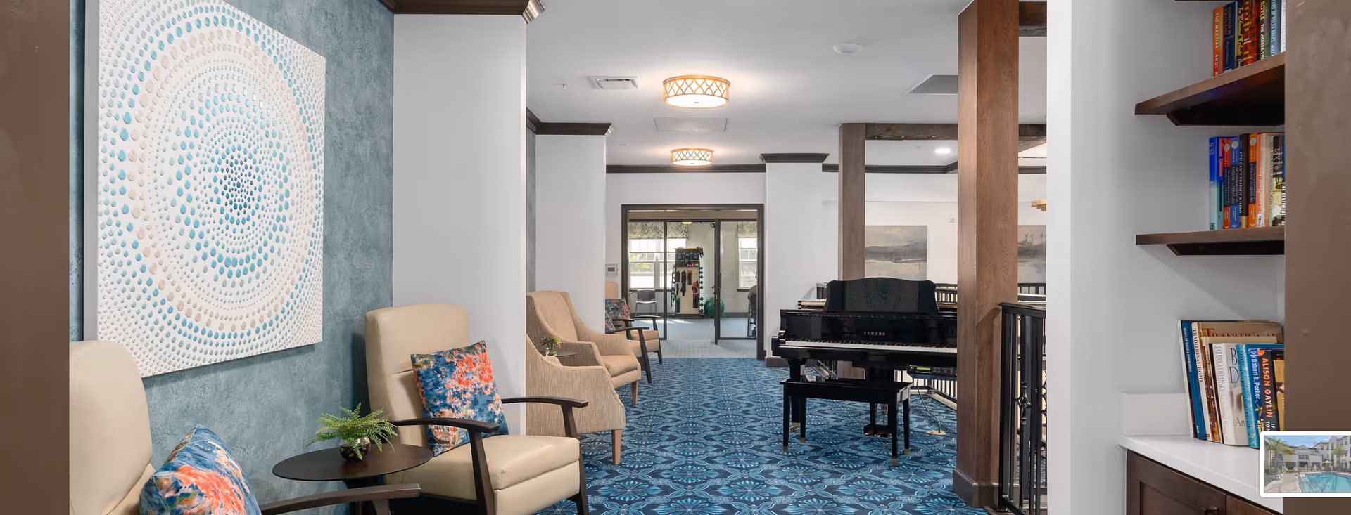 Interior view of a senior living facility lounge area with comfortable armchairs featuring colorful cushions, a small round table with a plant, a blue patterned carpet, a black Yamaha piano, wooden pillars, and bookshelves filled with books. The walls are decorated with abstract artwork, and there is a glass door leading to another room in the background.