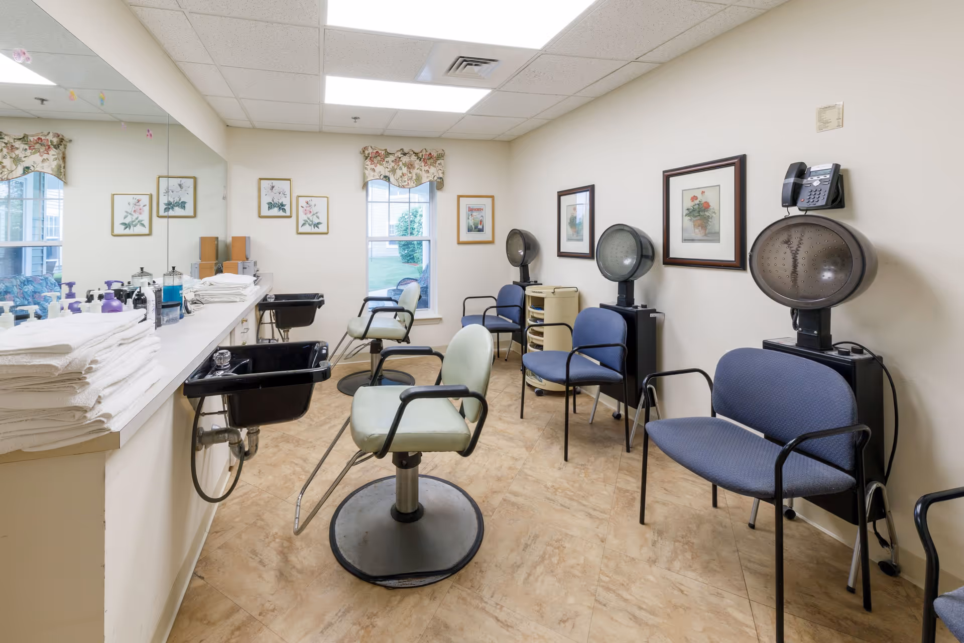 Interior view of a salon area in an assisted living facility with salon chairs, hair dryers, a sink, and a counter with towels and hair care products. The room has light-colored walls, framed floral artwork, and windows with floral valances.