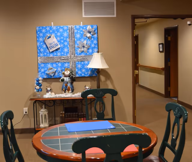 A cozy interior room with a round wooden table and four green chairs. On the wall behind the table is a large blue decorative piece with silver ribbons and snowflake patterns, along with winter-themed decorations including a snowman figurine, a lamp, and other small ornaments. There is an open doorway leading to a hallway with beige walls and carpeted floors.