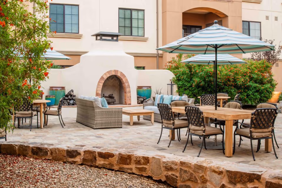A sunny outdoor courtyard with wooden tables, cushioned chairs, striped umbrellas, a wicker sofa, and a stucco fireplace in front of the building.