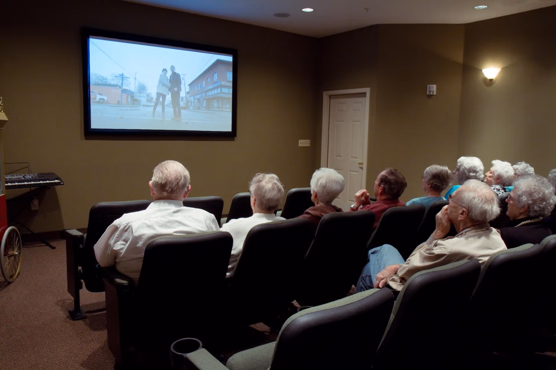 A group of elderly people seated in a small theater room watching a movie projected on a screen. The room has beige walls, theater-style chairs, and a keyboard on the left side.