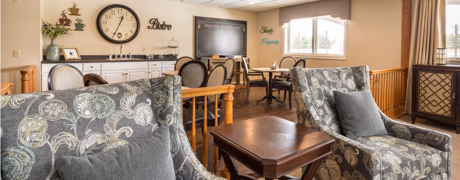 A cozy common area in a senior living facility featuring two patterned armchairs with gray cushions, a wooden side table between them, and a dining area in the background with round tables and chairs. The wall has a large clock, decorative coffee cup wall art, a chalkboard labeled 'Branch Happenings,' and the words 'Family' and 'Happiness' displayed. Large windows with curtains allow natural light into the room.