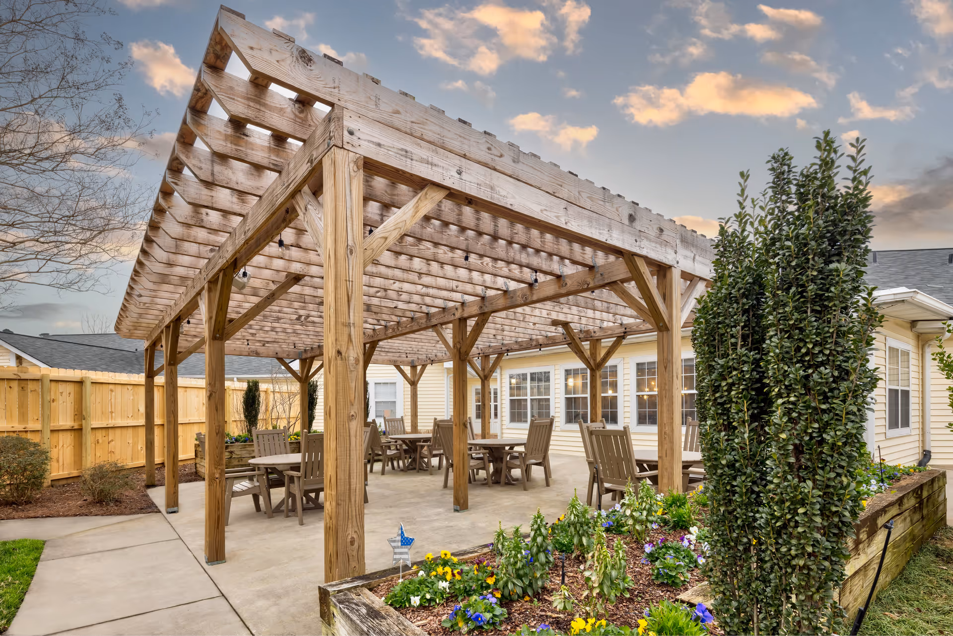 Outdoor patio area at TerraBella Cramer Mountain featuring a wooden pergola with several tables and chairs underneath. The patio is surrounded by a wooden fence, flower beds with colorful flowers, and greenery. The building with multiple windows is visible in the background under a partly cloudy sky at sunset.