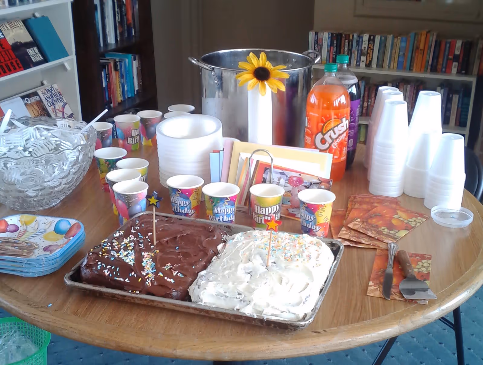 A round wooden table set up for a birthday celebration with two cakes, one with chocolate frosting and the other with white frosting and sprinkles, surrounded by colorful 'Happy Birthday' paper cups, plastic utensils in a glass bowl, stacks of paper plates and white disposable cups, two bottles of soda, and a large metal container with a yellow artificial sunflower attached. Bookshelves filled with books are visible in the background.