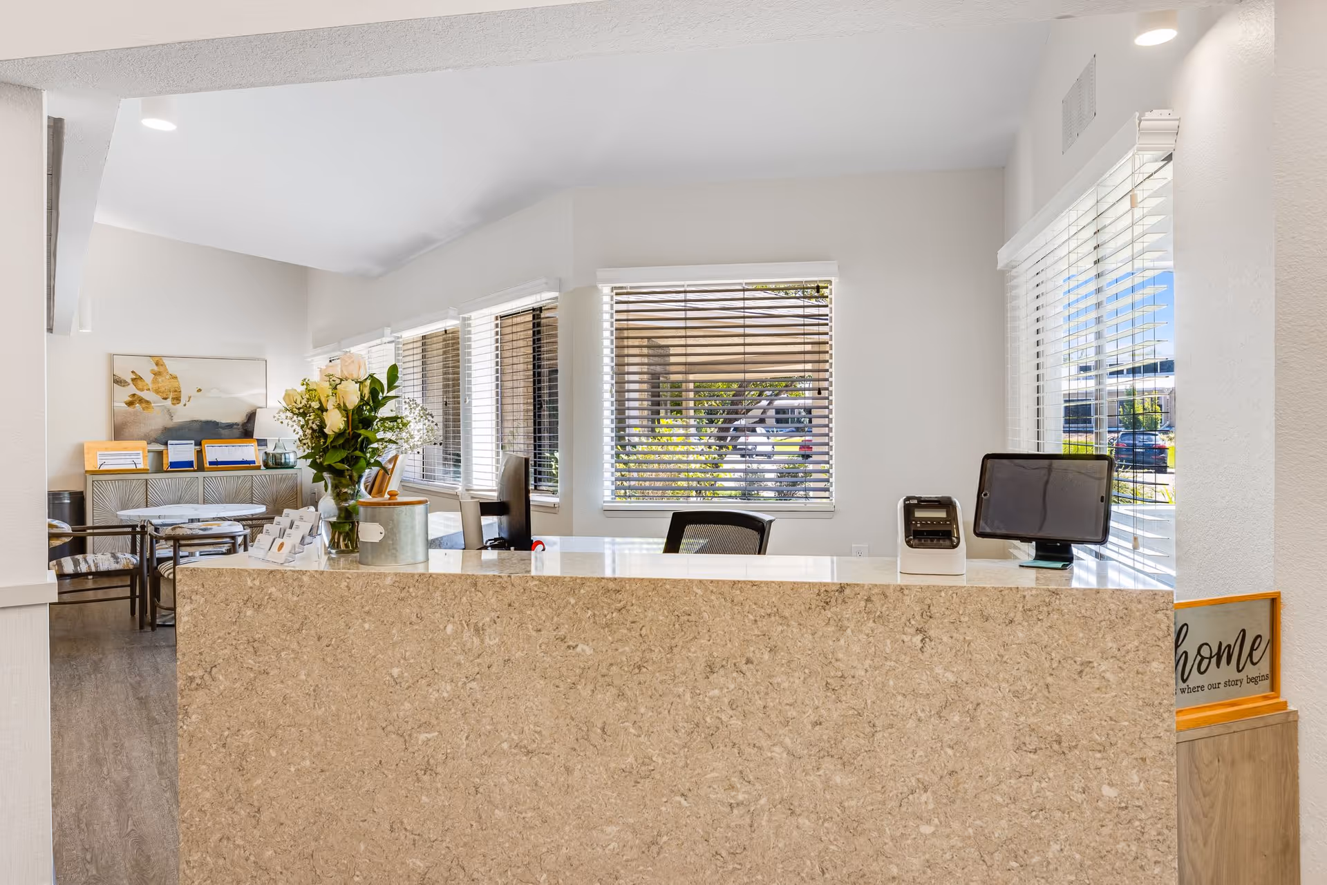 Reception area of a senior living facility with a beige stone front desk, a vase of white roses, a tablet device, and a small sign that says 'home where our story begins'. Behind the desk are windows with blinds and chairs, and a seating area with tables and chairs is visible to the left.