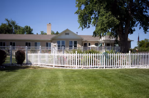 Single-story assisted living building seen behind a white picket fence with a manicured lawn and large trees.