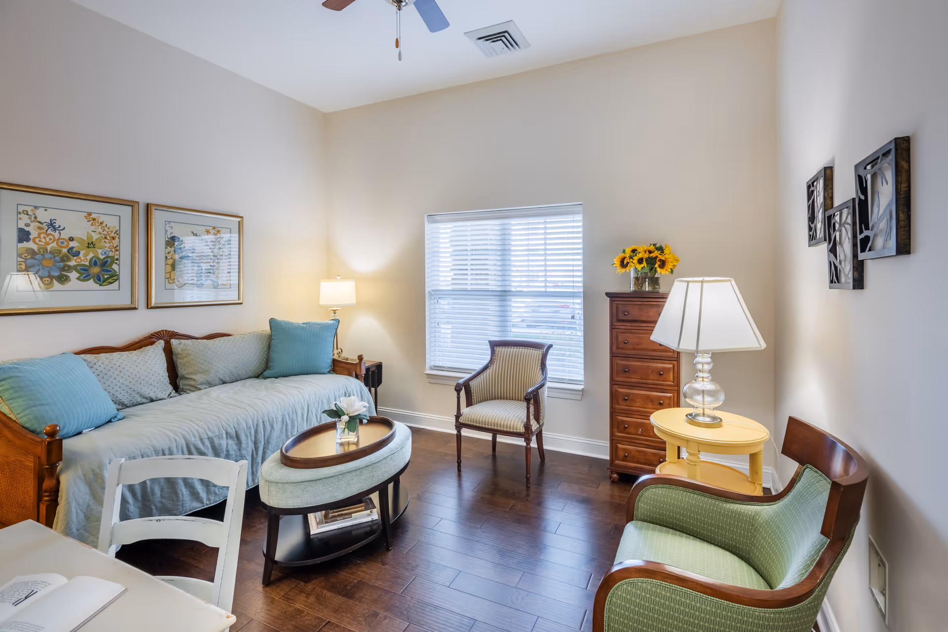 A cozy living room with a wooden daybed covered in light blue cushions and pillows, a small oval coffee table with a flower vase, two armchairs, a wooden chest of drawers with a vase of sunflowers on top, a yellow side table with a lamp, and framed artwork on the walls. The room has a window with blinds and wooden flooring.