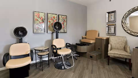 Interior of a salon area in a senior living facility with two beige salon chairs with black armrests and hair dryers, a small round table between them, a beige pedicure chair with a foot bath, and a beige patterned armchair. The walls are decorated with floral artwork and mirrors, and the floor has wood-like flooring.